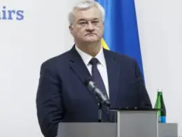 Ukrainian Foreign Minister Andrii Sybiha stands behind a lectern with a microphone and a Ukrainian flag in the background.